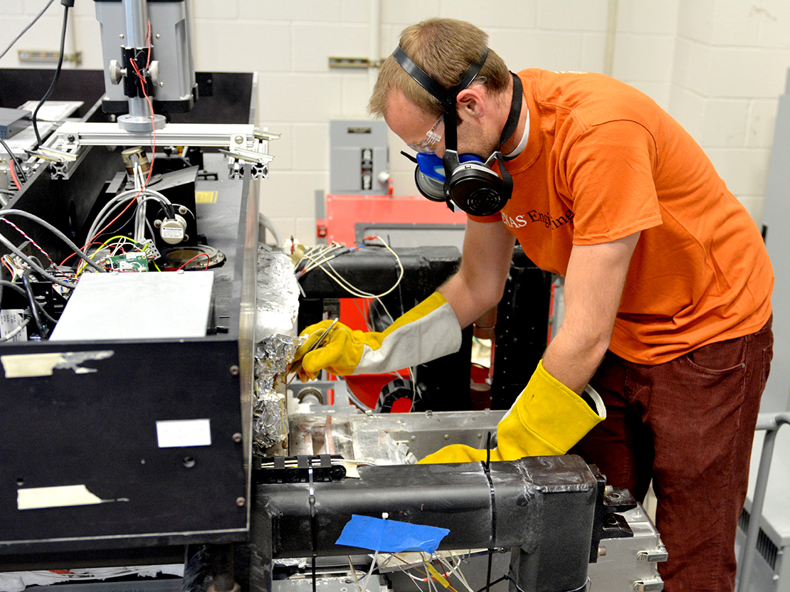Researcher wearing PPE handling small instruments next to large machine in lab.